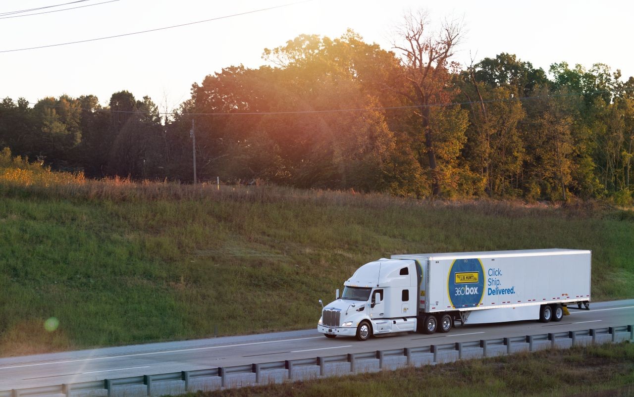 A white semi‑truck hauling a branded 360box trailer drives along a highway at sunset, with a grassy hillside and trees in the background.