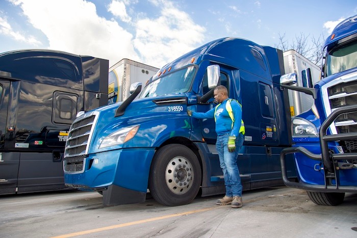 Blue semi-truck parked at a lot with other trucks, person in safety gear standing beside it.