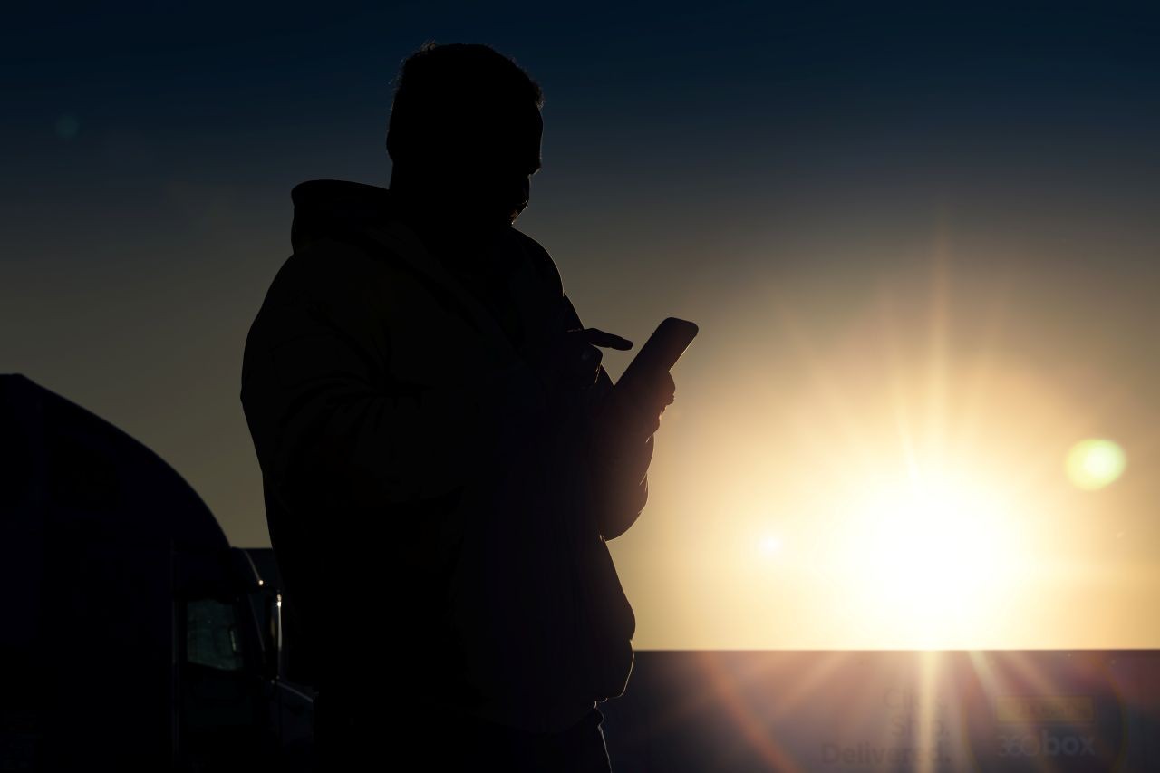 Owner operator in silhouette checking his mobile app for updates while the sun sets in front of him