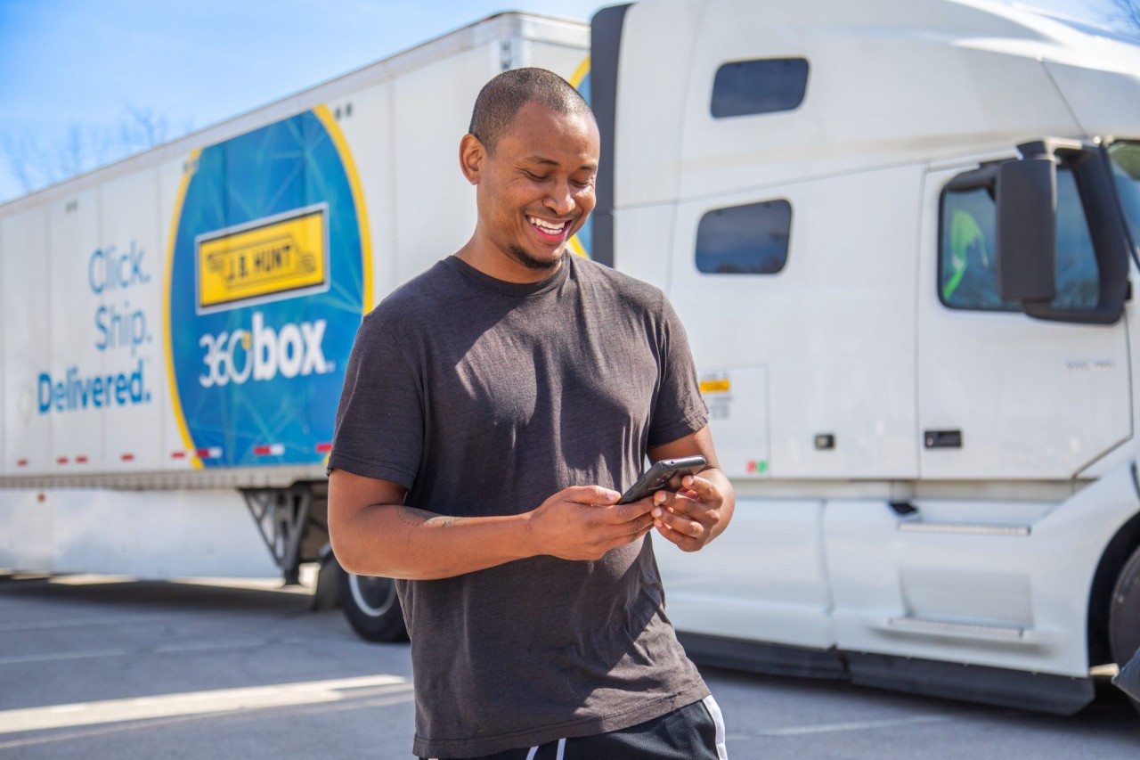 J.B. Hunt owner operator stands in front of a J.B. Hunt 360 box truck while smiling and using their phone.
