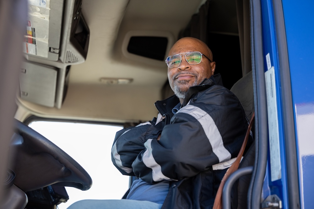 An experienced African American J.B. Hunt owner operator truck driver, wearing safety glasses, smiles from his cab. This image is featured on the J.B. Hunt Owner Operator hiring website's Frequently Asked Questions section, symbolizing a trusted resource.
